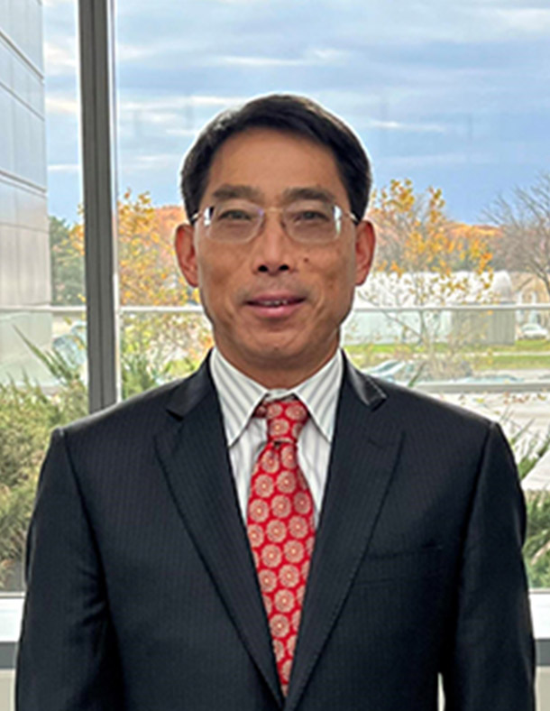 Shaomeng Wang standing indoors in front of large windows that look out on ground floor buildings.