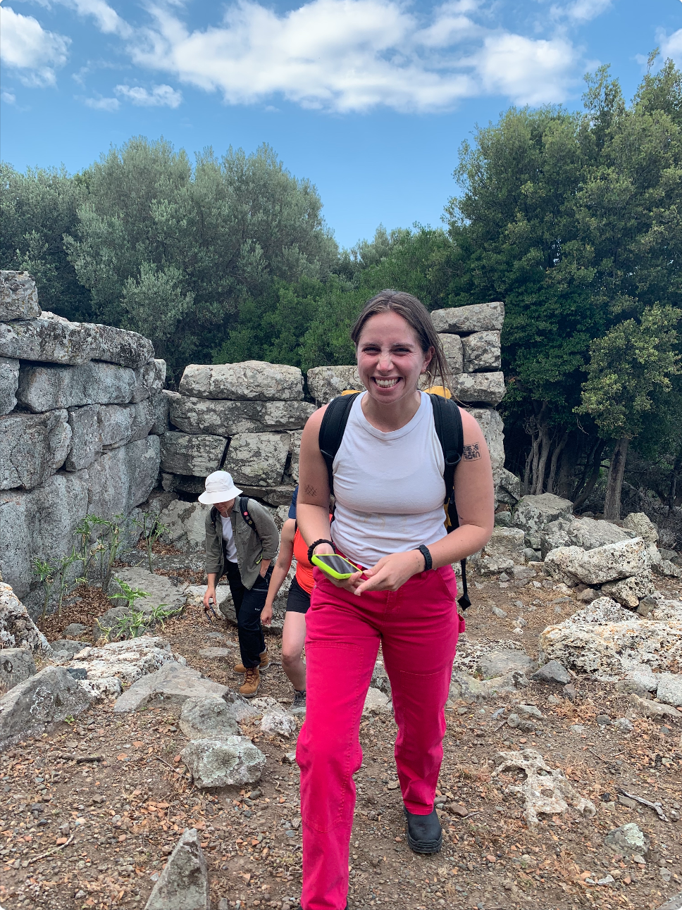 A brunette woman in pink pants and white tank grins excitedly while hiking up rocky terrain, a ruined wall behind her.