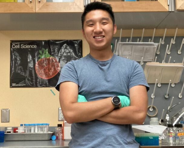 Biology major Ethan Lane stands with his arms crossed smiling for the camera. He's wearing medical gloves and is in a research lab.